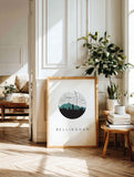 a framed poster of a city skyline with the word "Bellingham" on it, placed on a wooden floor in a room with a couch, a plant, and some books.